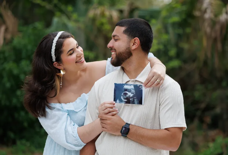 A couple joyfully holds an ultrasound image outdoors, celebrating their upcoming arrival, surrounded by greenery.