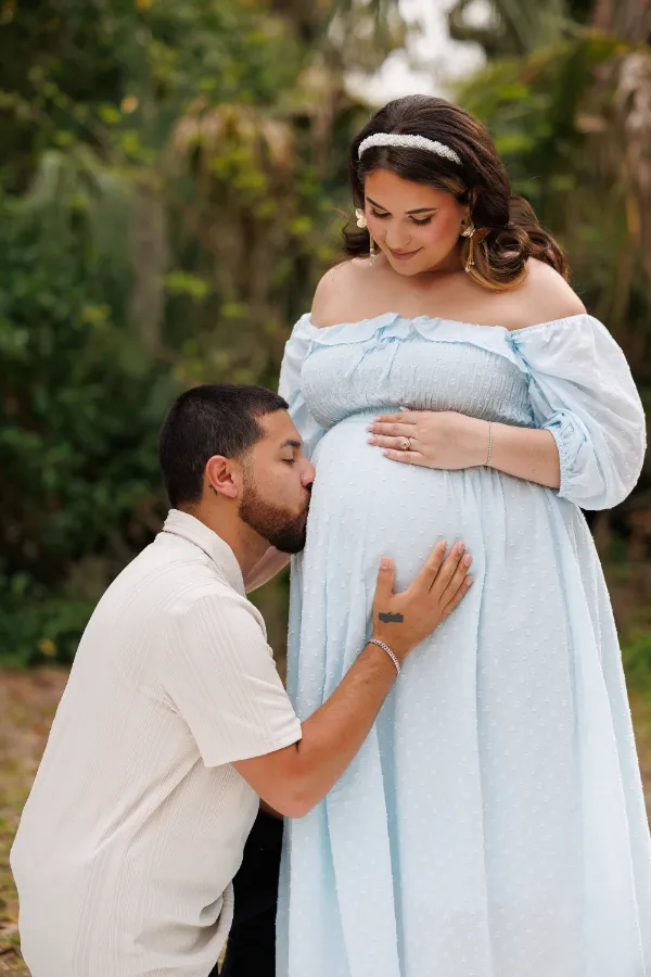 A man kneels in front of a woman wearing a light blue dress, gently touching her pregnant belly in a lush outdoor setting.