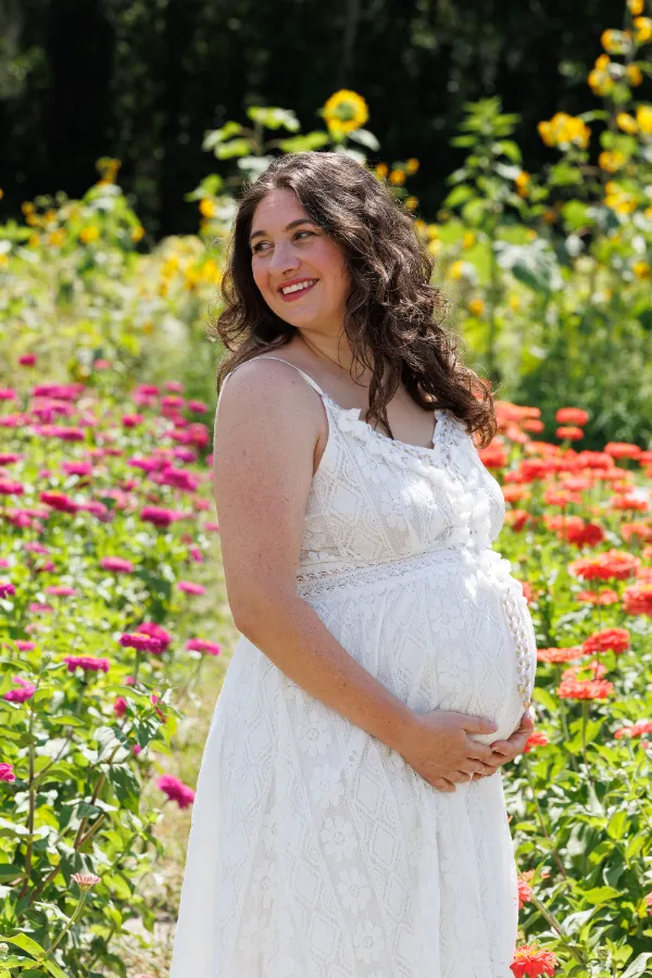 A pregnant woman in a white dress stands among vibrant flowers, cradling her belly in a sunny, blooming garden.