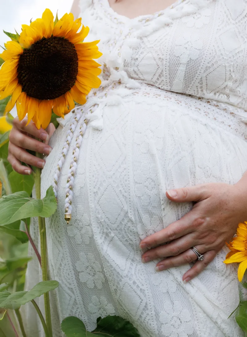 LO1A4917 A pregnant woman in a white lace dress cradles her baby bump while holding a vibrant sunflower surrounded by greenery.