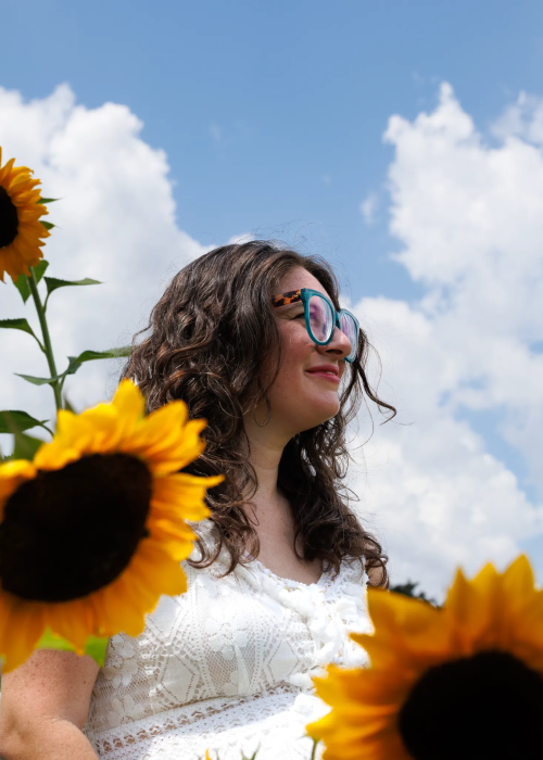 A woman stands amidst vibrant sunflowers, with a bright blue sky and fluffy clouds in the background, radiating summer joy