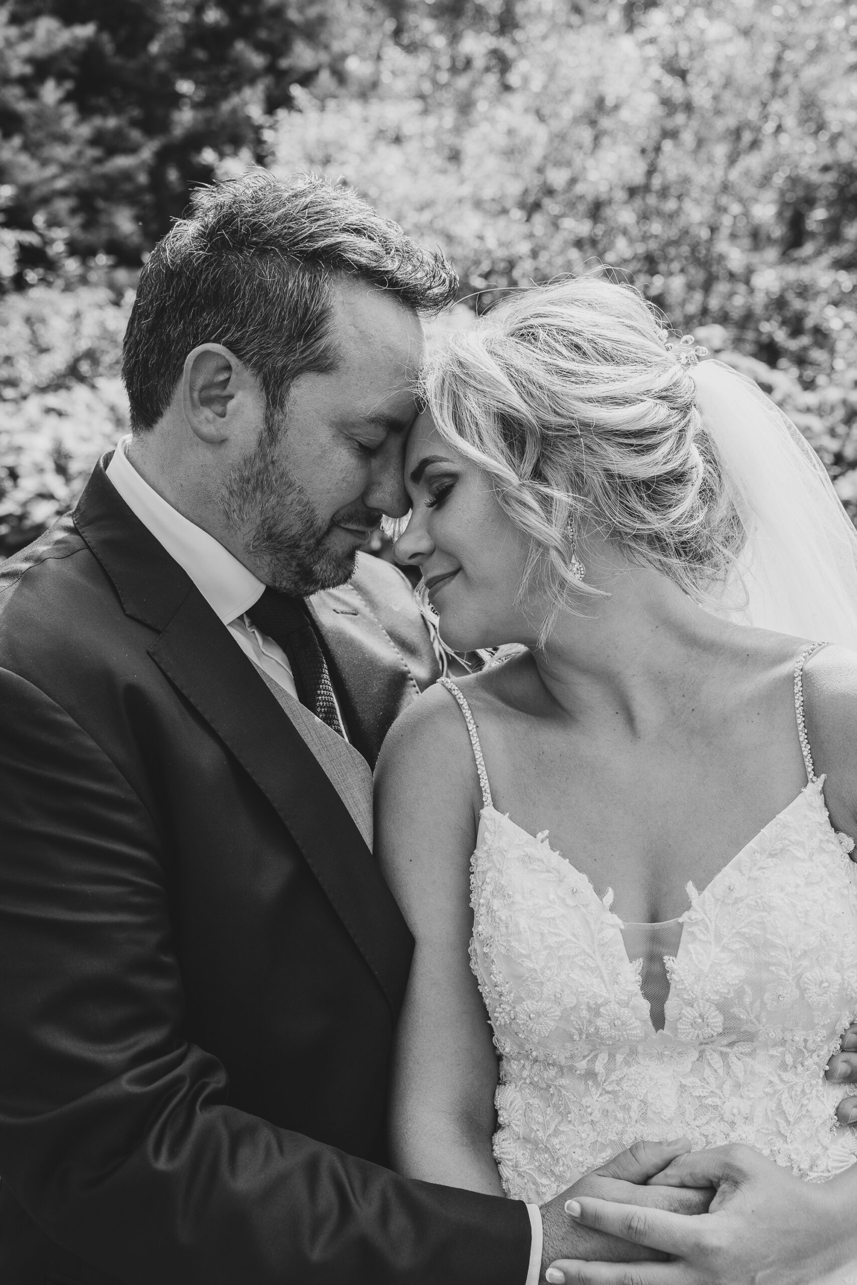 7N4A7797-2 A pregnant woman in a white lace dress cradles her baby bump while holding a vibrant sunflower surrounded by greenery.